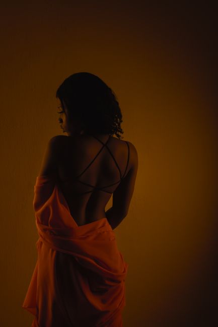 Back view of slim African American female with dark hair in stylish clothes standing in studio against dark orange background