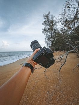 Crop anonymous traveler demonstrating modern professional photo camera on sandy beach near wavy sea against cloudy sky