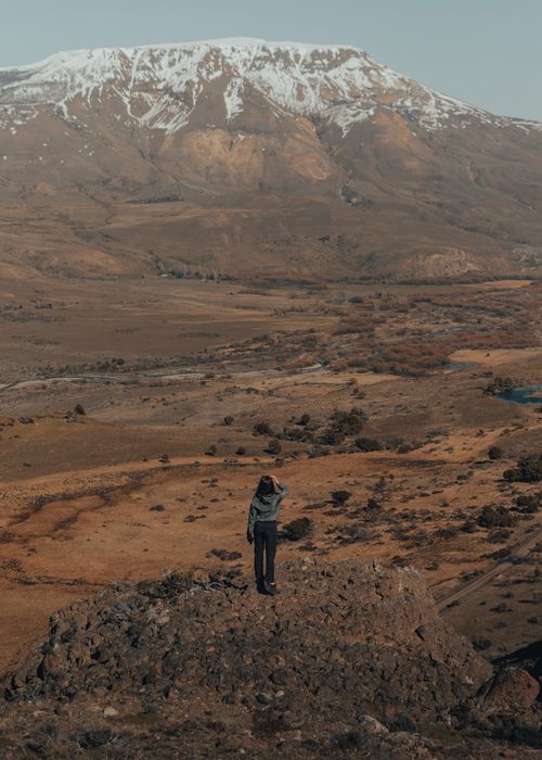 Free A lone woman stands on rocky terrain gazing at a snow-capped mountain, capturing the essence of adventure and nature. Stock Photo