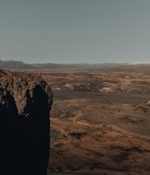 A lone figure stands on a cliff overlooking a vast desert landscape under a clear sky.