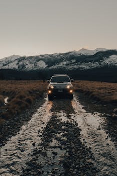 SUV navigating a rocky dirt road with snow-capped mountains in the background under a moody sky.