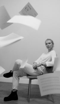 Striking black and white portrait of a woman seated, surrounded by flying papers, indoors.