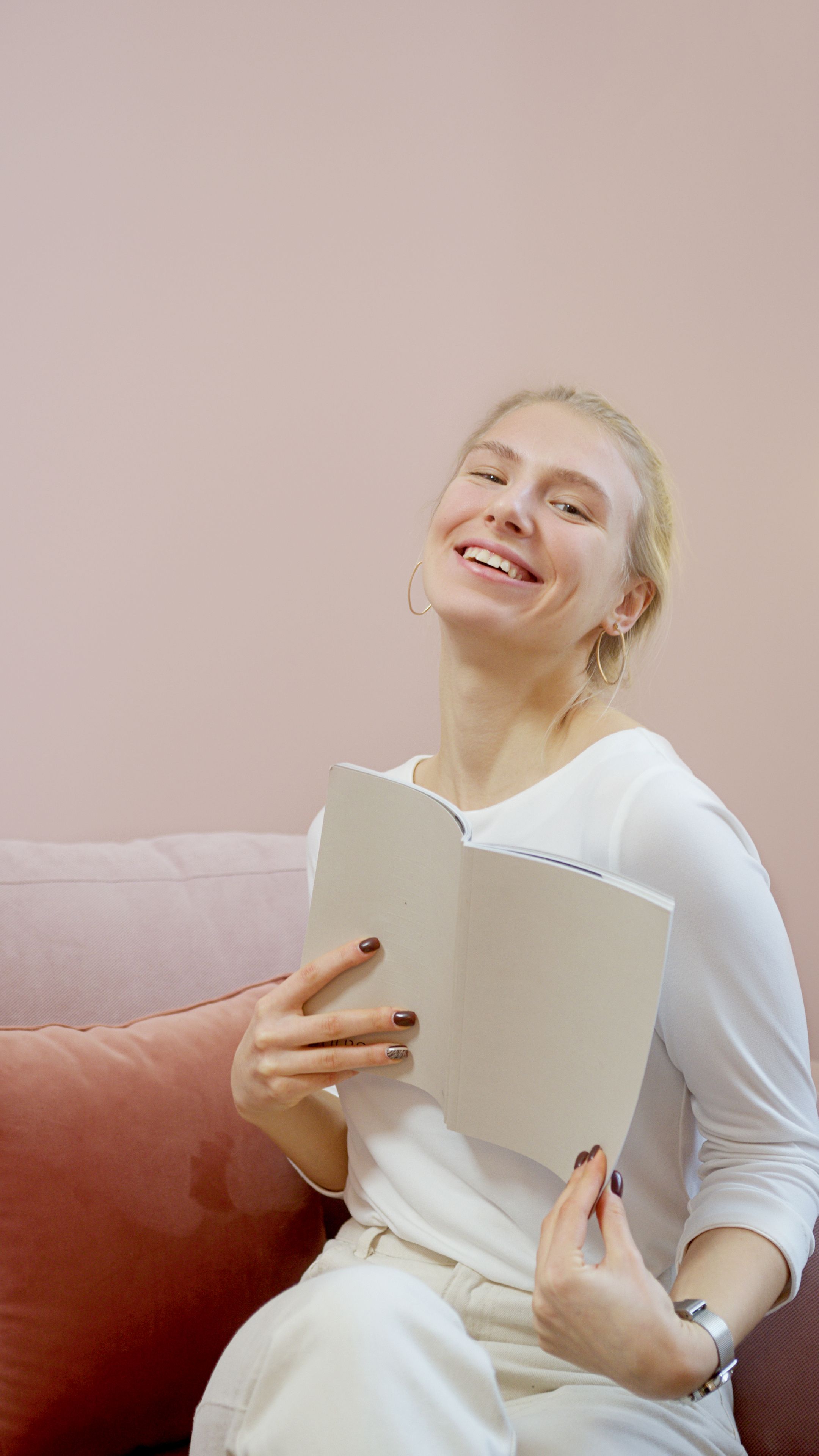 Cheerful woman in white holding a book on a pastel sofa, exuding warmth and joy.
