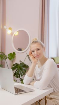 Young woman with blond hair sitting at a table with a laptop, smiling in a modern indoor setting.
