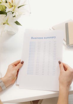 Hands holding a business summary document at a desk with flowers, papers, and a calm interior.