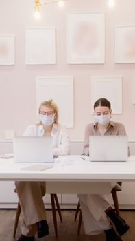 Two women working in an office setting, both wearing face masks and using laptops.