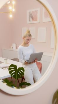 Caucasian woman working on a laptop from home office, reflecting in a mirror. Modern and stylish interior.