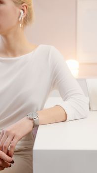 Sophisticated woman wearing earbuds and wristwatch, sitting elegantly indoors with modern lighting.