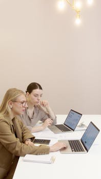 Two professional women collaborating on laptops in a modern office setting.