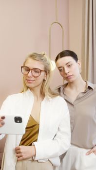 Two fashionable women posing together and taking a selfie with a smartphone indoors.
