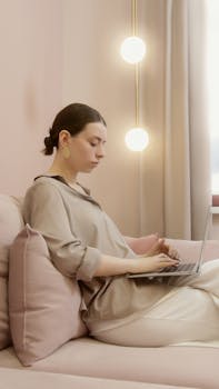 A focused woman typing on her laptop while sitting on a comfortable sofa in a calm, indoor setting.