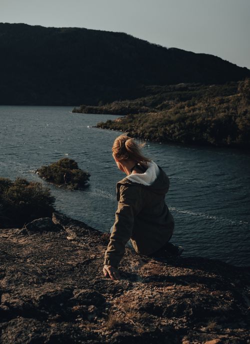 Free Woman sitting on a cliff edge overlooking a serene lake and mountains under clear skies. Stock Photo