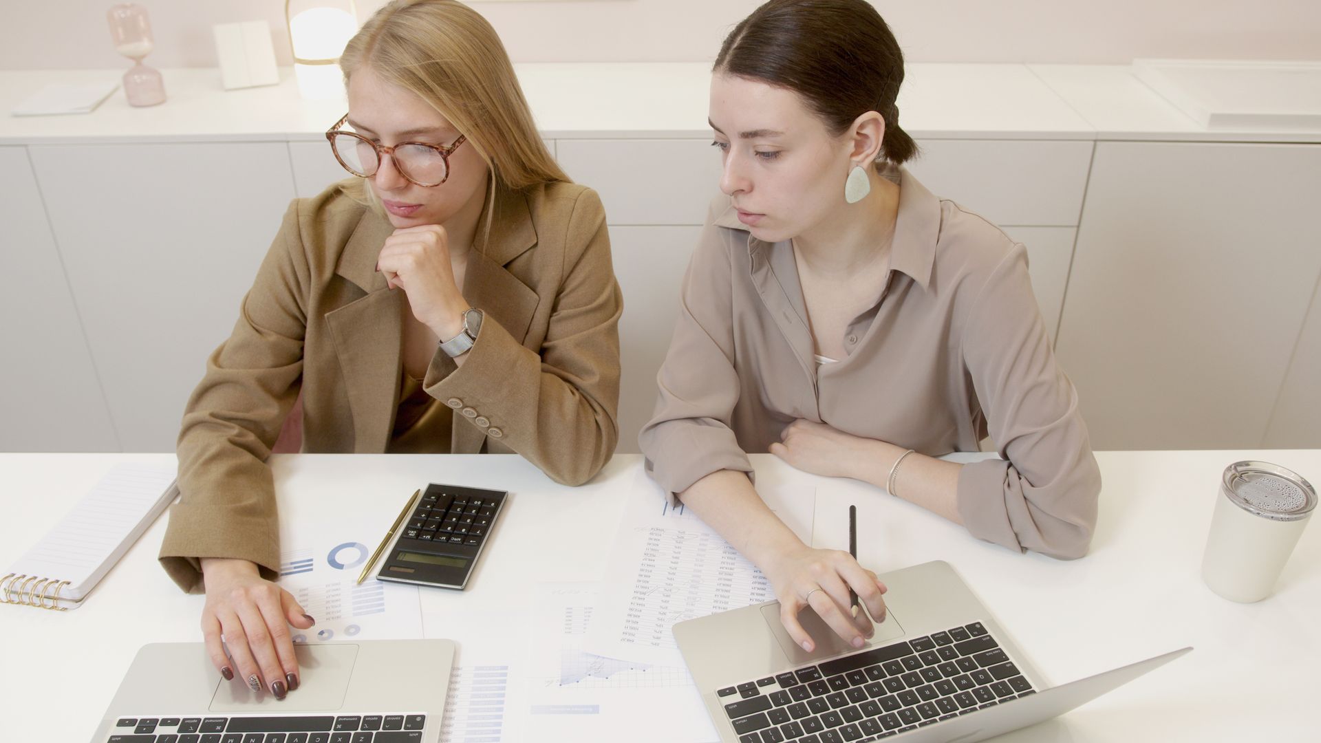 Two women working together with laptops and documents in a contemporary office setting.