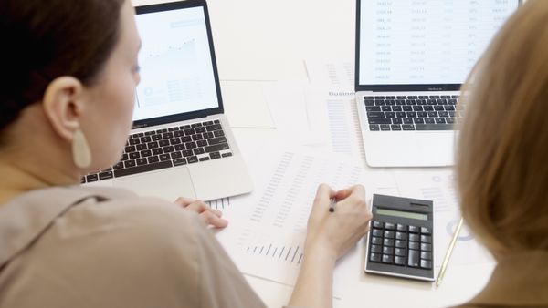 Two women at a white desk with a laptop showing figures and a calculator beside printed sheets