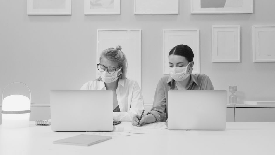 Dos mujeres trabajando de manera colaborativa con mascarillas en un entorno de oficina moderna.