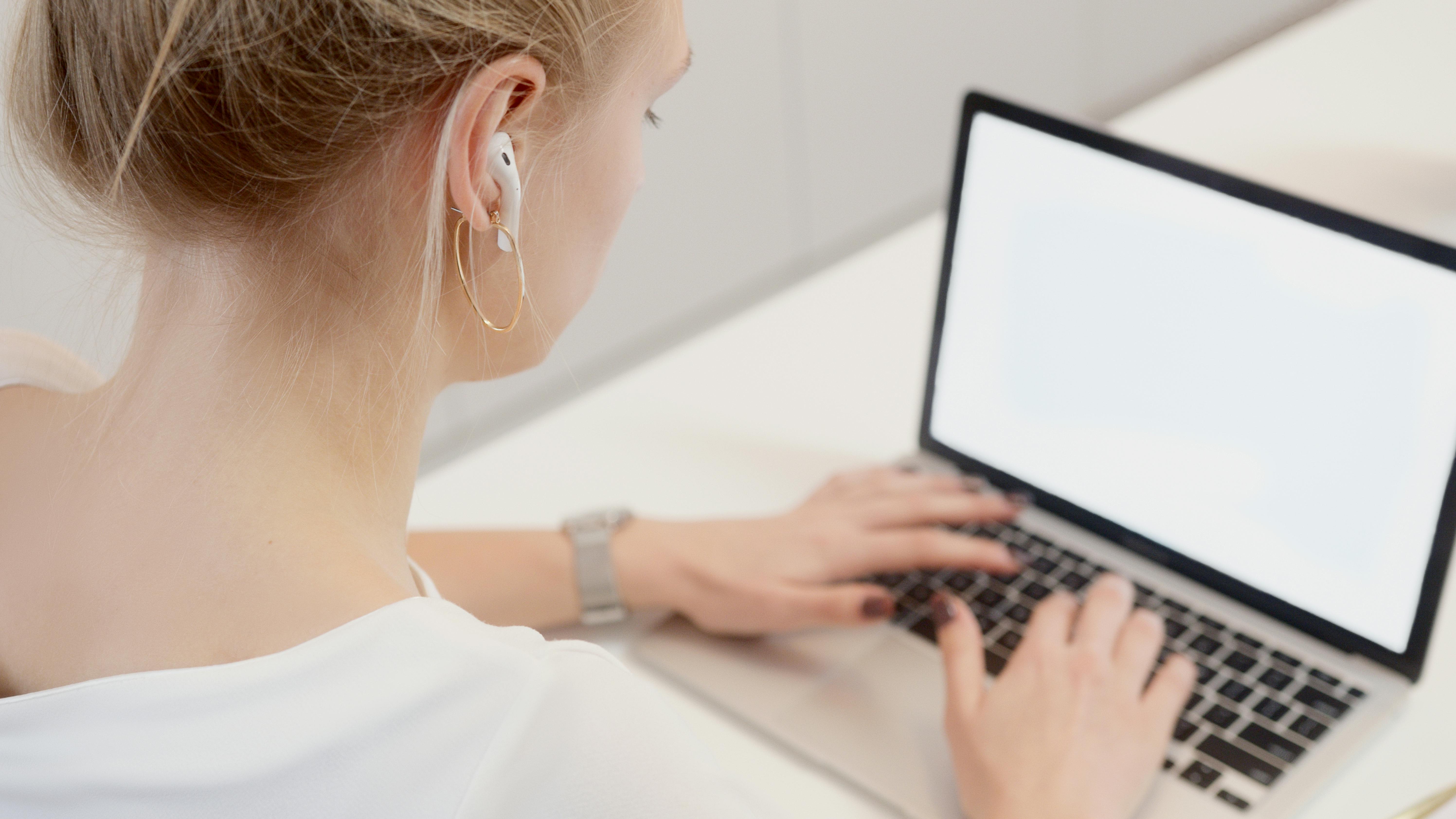 wireless earbuds with long battery life uk - Adult woman typing on a laptop while wearing wireless earbuds in a bright indoor setting.