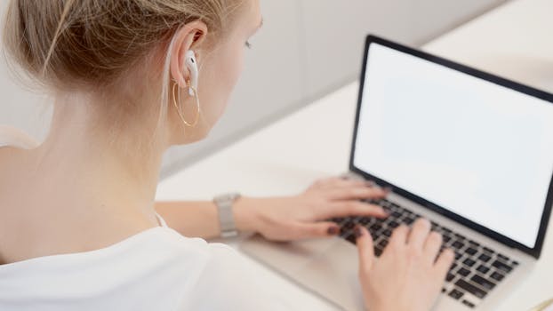 Adult woman typing on a laptop while wearing wireless earbuds in a bright indoor setting.