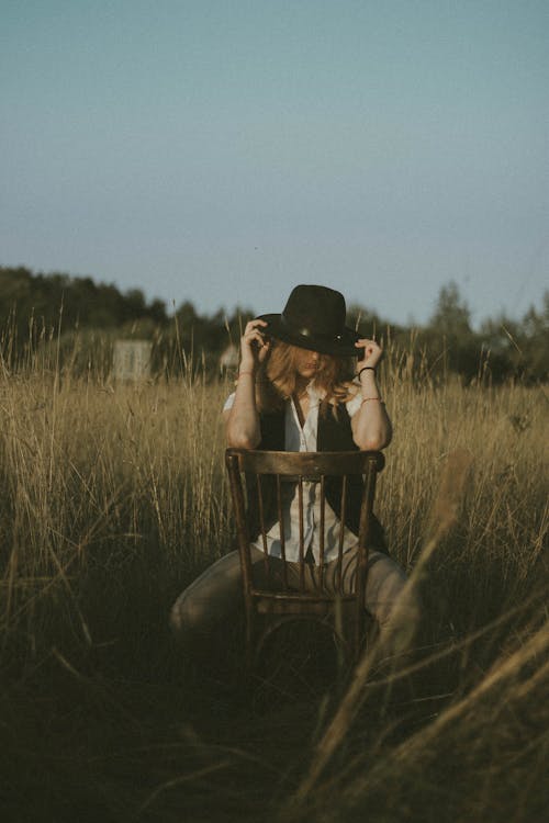 Unrecognizable stylish woman with hat on head leaning elbows on back of chair while sitting in field on wooden chair