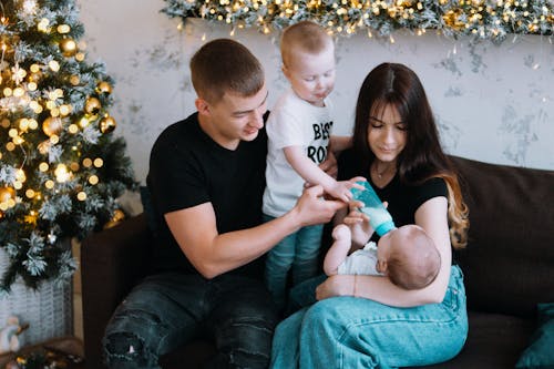Caring parents and elder son feeding baby together sitting on couch near Christmas tree and garland