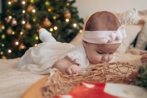 Toddler girl in white dress lying on comfortable bed near pillows and decorative objects in light bedroom with Christmas tree with baubles and garland at home