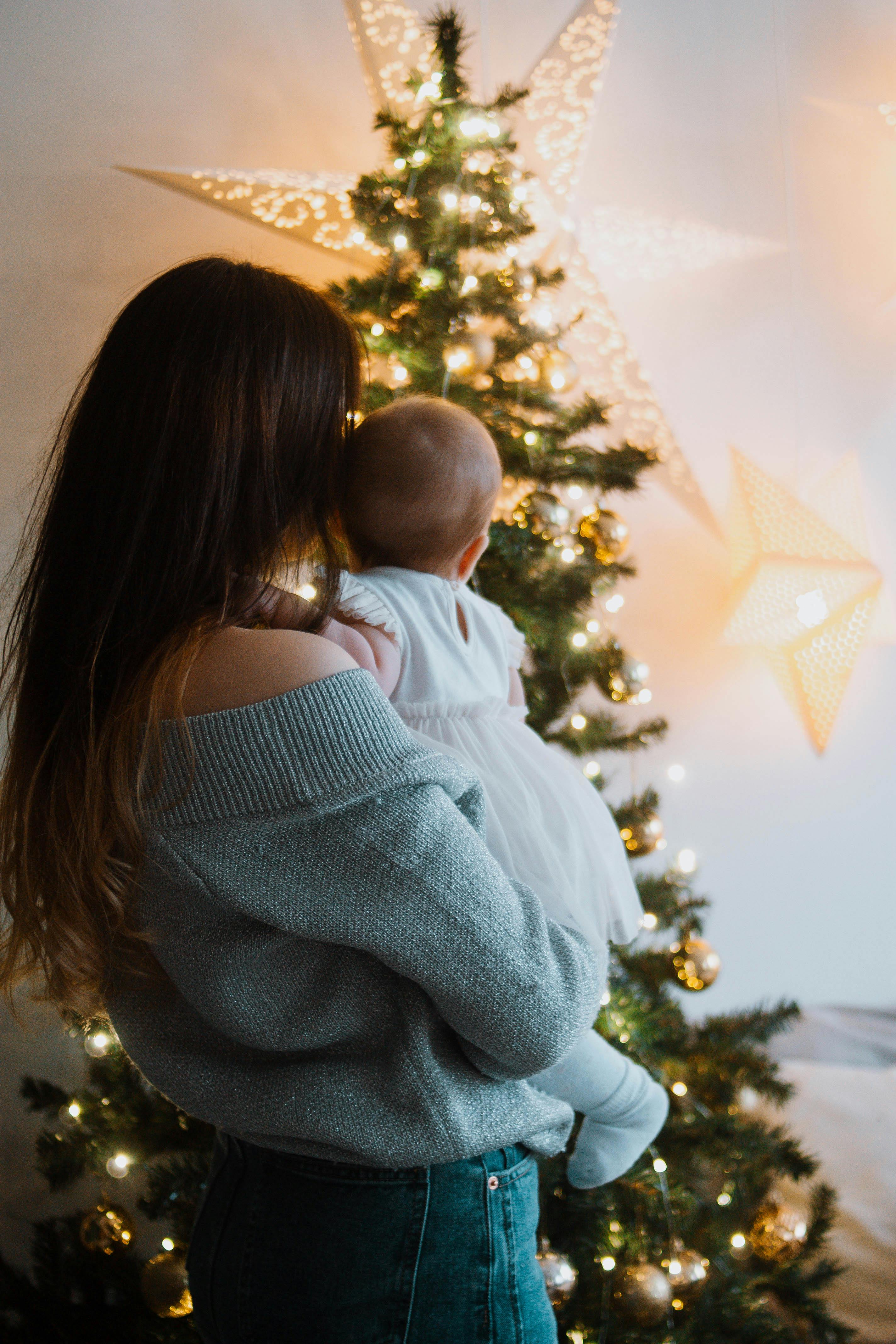 Back view of anonymous mother standing with little baby on hands and looking at Christmas tree with festive decorations and garlands at home