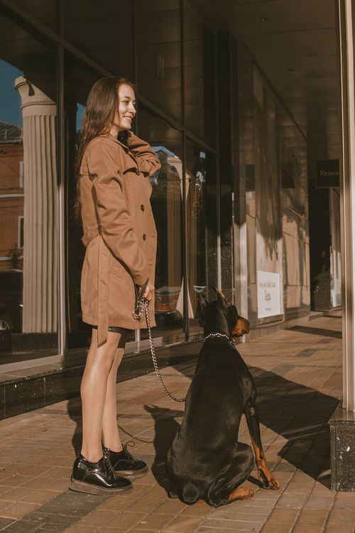 Free A woman in a brown coat stands with a Doberman on a sunny urban street. Stock Photo