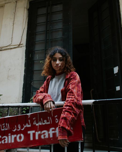 Sad young ethnic lady in casual clothes standing near metal railing with signboard with inscription in city street near building in daytime while looking at camera