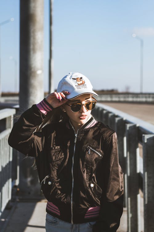 Young stylish female wearing stylish outfit and sunglasses standing with hand on head near metal railing and looking away