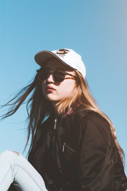 Low angle of serious female in trendy sunglasses and cap with flowing hair against blue sky