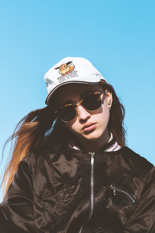 Low angle of serious female in stylish sunglasses and cap with flying hair against blue cloudless sky