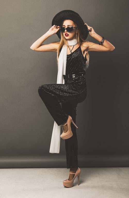 Full body of elegant female in elegant outfit with sunglasses standing with leg raised and hands on head in studio