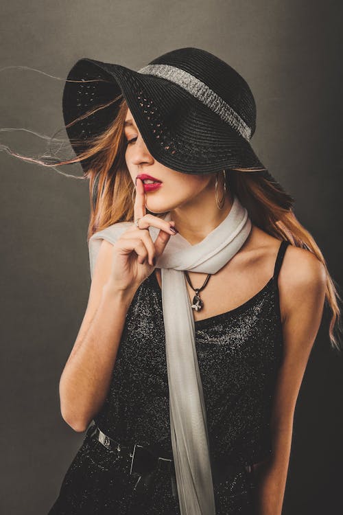 Female with long flying hair wearing trendy hat and scarf touching lips with forefinger while making shh gesture in studio