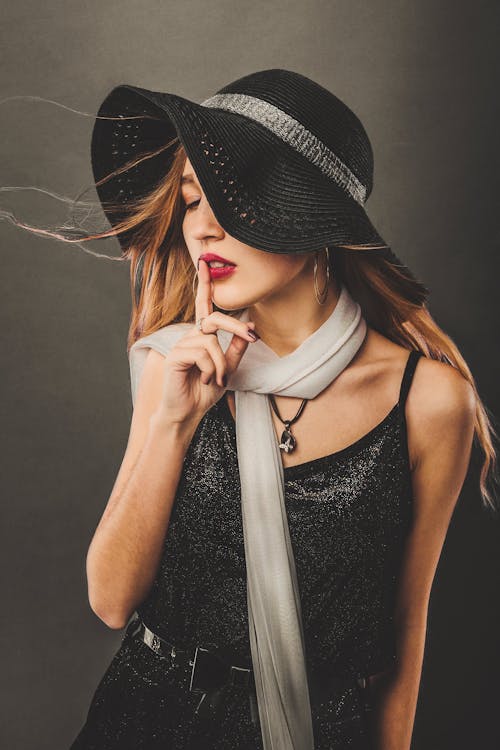 Free Female with long flying hair wearing trendy hat and scarf touching lips with forefinger while making shh gesture in studio Stock Photo