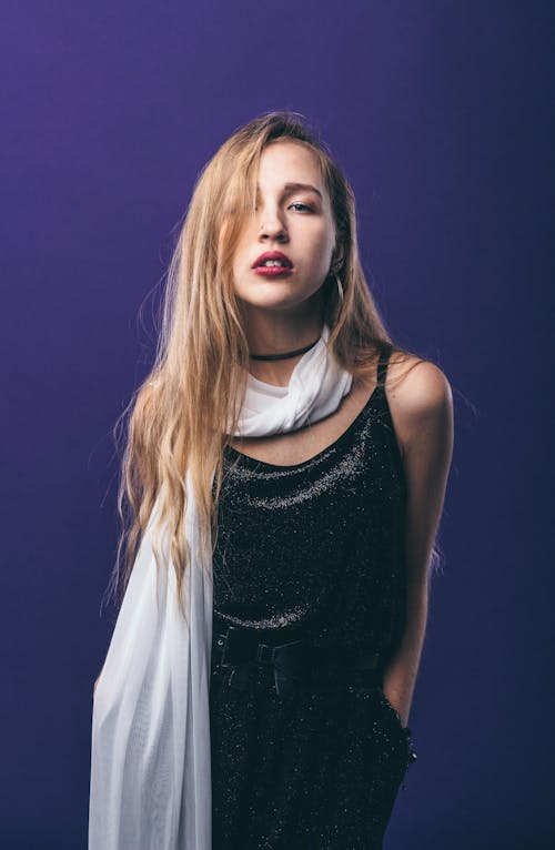 Provocative female wearing stylish outfit with long hair standing with hands in pockets against blue background and looking at camera