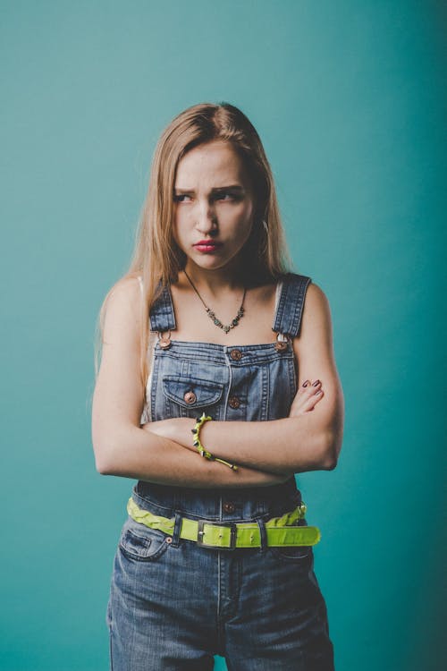 Displeased female model in casual denim overall looking away while standing against blue background with crossed arms and raised shoulders