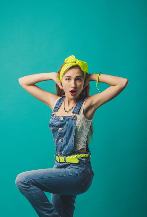 Astonished young female hipster wearing denim overall and bright neon accessories with armes raised behind head and raised knee and looking at camera