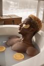Thoughtful naked African American man with blond hair and eye patches lying in bathtub with slices of orange in spa and looking away