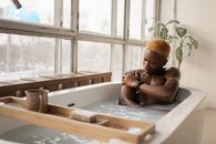 Young African American man with dyed hair and accessory sitting in bathtub full of water in light room with shabby window frames