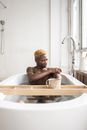 African American man sitting in bathtub with cup of coffee on tray and scrubbing arms with sponge and soap