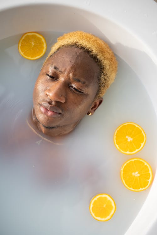 From above of Black man with colorful hair looking away while relaxing in bathtub full of water with slices of fresh orange