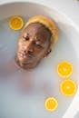 From above of Black man with colorful hair looking away while relaxing in bathtub full of water with slices of fresh orange