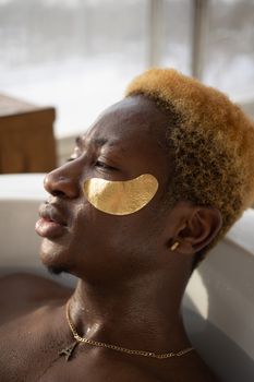 Side view of pensive African American man with dyed hair lying in tub with golden eye patches while relaxing in bathroom during skincare procedure