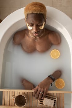 Top view of peaceful African American male with eye patches lying in bathtub with milk water and wooden tray with cup during spa procedure