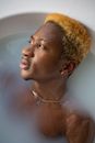From above of dreamy African American male with dyed hair looking away while relaxing in bathtub with milk water during daily routine