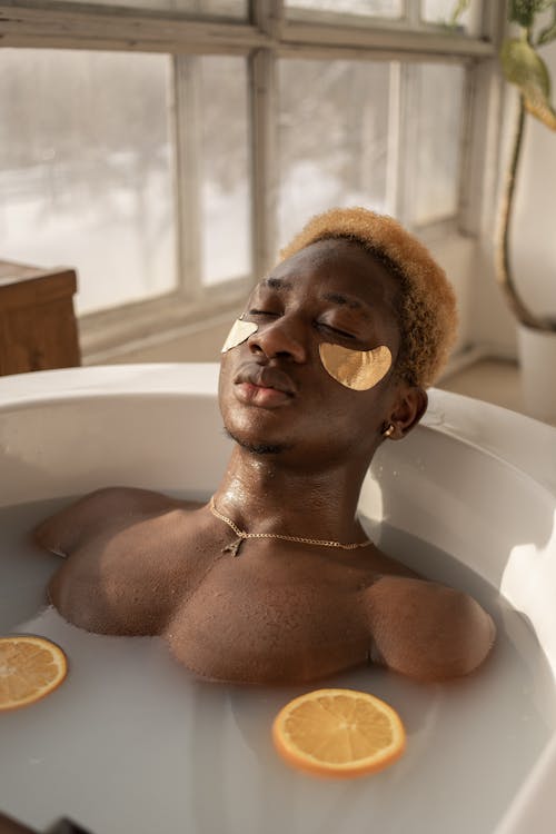 Relaxed African American male with dyed hair lying in bath with milk water and orange slices during spa procedure