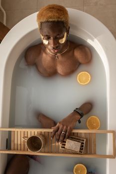 Top view of calm African American male with dyed hair lying in bathtub with orange slices and wooden tray with mug during skincare routine