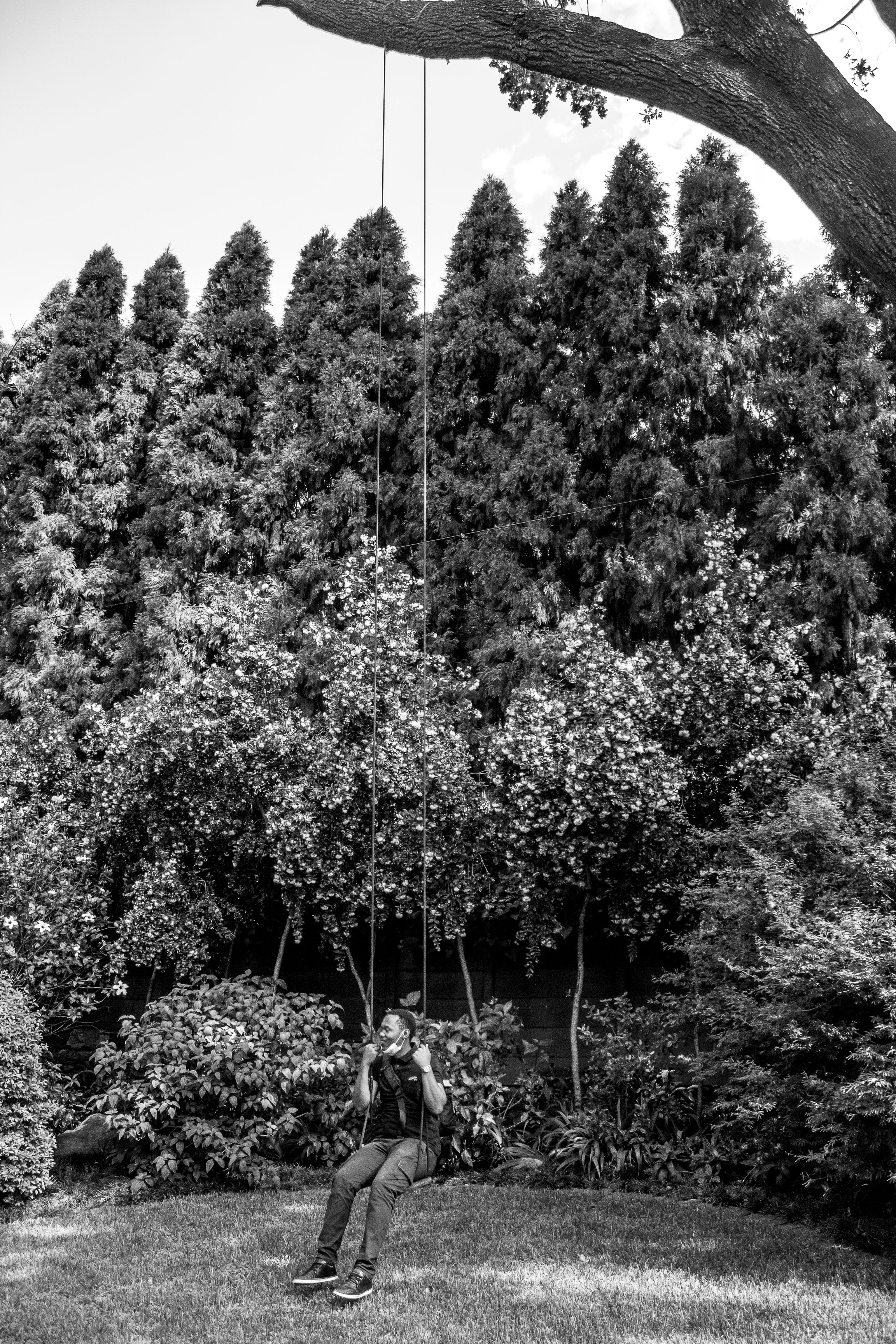 Monochrome image of a man sitting on a swing under a tree in a garden.