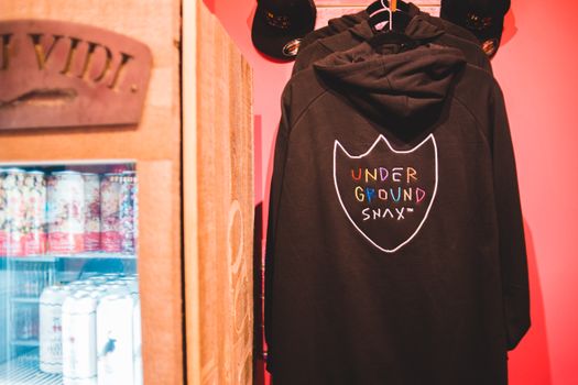 Stylish sweatshirts with hoods between caps against refrigerator with cans of beverages for sale in shop