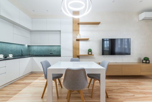 Modern flat interior with table with chairs next to shelves and TV on wall near open kitchen with cupboards