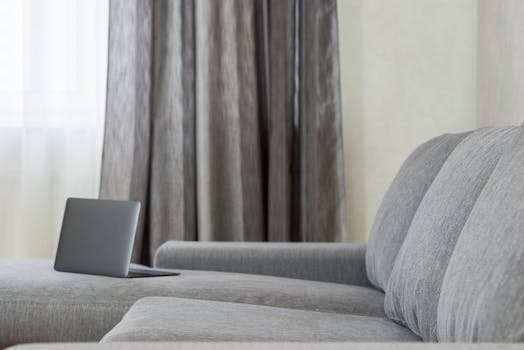 Cozy and bright modern living room with a laptop on a grey sofa, perfect for remote work.